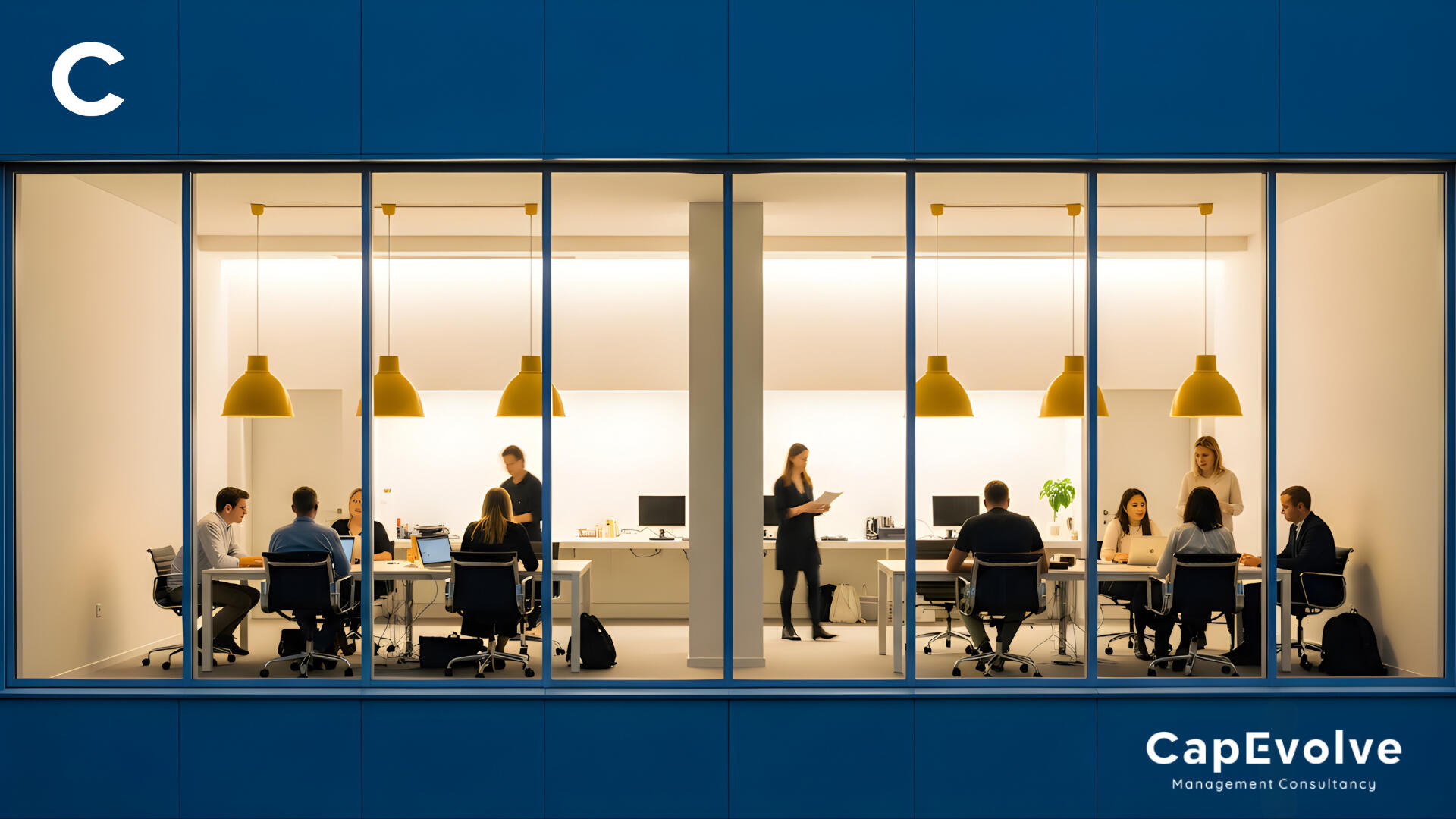 CapEvolve Management Consultancy office viewed through large windows. Inside, diverse team members collaborate at desks under warm yellow pendant lights. One woman stands reviewing documents, others work on laptops or discuss in small groups. Clean, modern